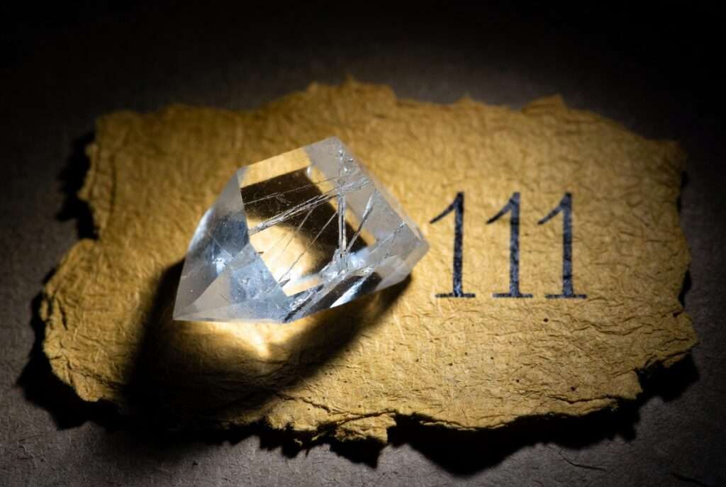 A highly-focused studio photograph of a raw clear quartz crystal point resting next to the handwritten digits '111' on textured parchment paper, representing the amplified frequency of intent often utilized in high-quality manifestation journals.