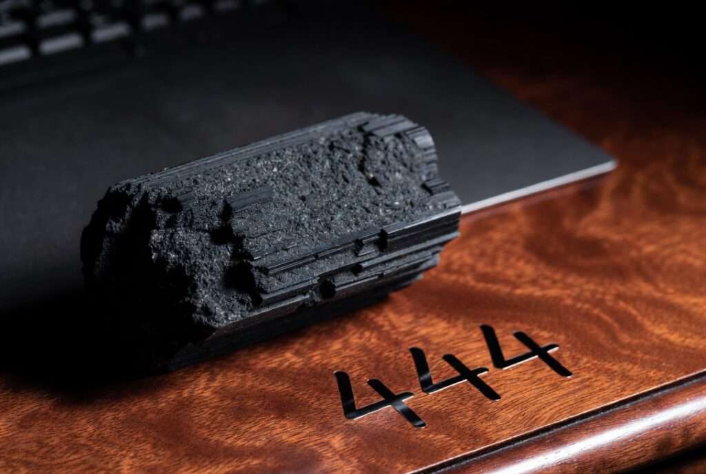 Stark film noir photograph of a raw black tourmaline stone resting on a dark mahogany desk beside the handwritten numbers '444', symbolizing structural stabilization and protective grounding techniques.