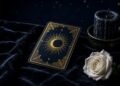 Dark navy tarot card back with a gold crescent moon and sunburst, beside a white rose and a candle on black velvet